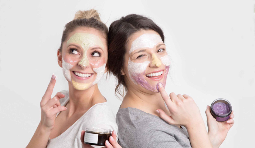 Две молоды женщины с нанесенными на лицо масками в технике мультмаскинг стоят спиной друг к другу и улыбаются Женщины используют маски для лица