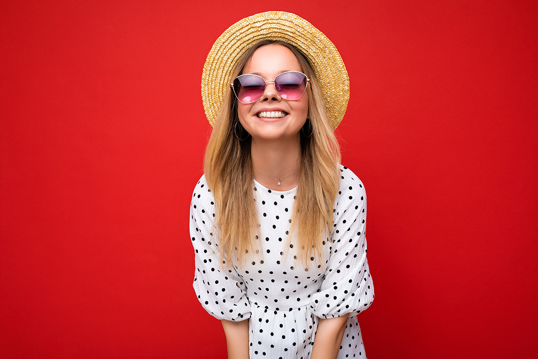 Девушка в белом платье в горошек, соломенной шляпе и солнечных очках улыбается Девушка в шляпе и в очках
